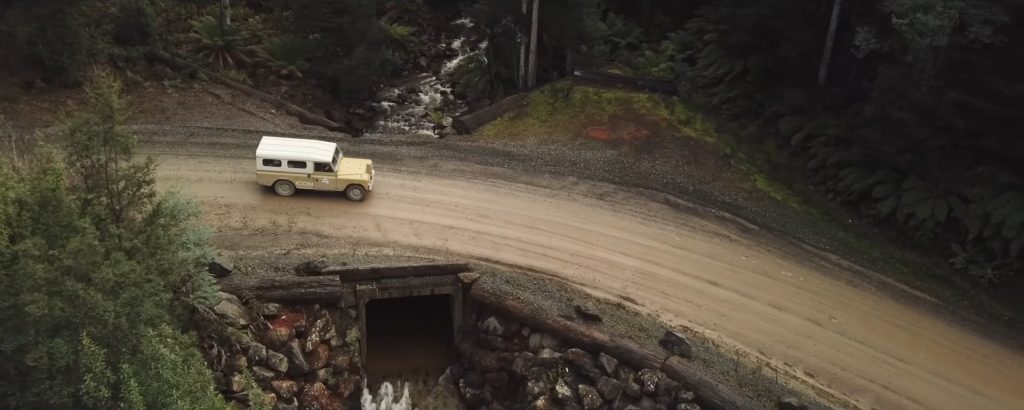 Ariel view, looking down at a Land Rover Series 3 in beige, with a white roof, driving left to right on a dirt track, crossing a culvert propped up by logs. The impression is it's in the middle of a forest.