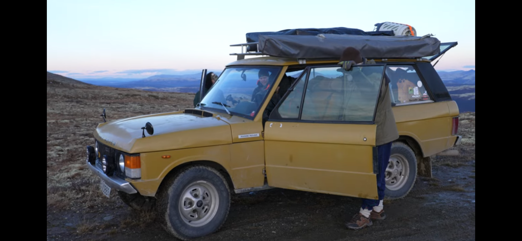 A 3 door classic Range Rover in mustard yellow, with a roof rack. A person is standing in the doorway of the car and there are mountains trailing down in the background.