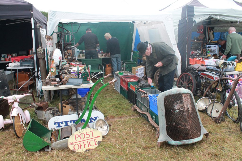 An assortment of old and vintage auto spares on a grass pitch. 4 men are in the frame and one, wearing a hat, is bending over looking through a box of spares.