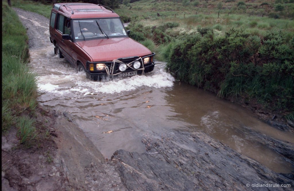 Red Land Rover Discovery 1 entering a smallish puddle. There is a bow wave at the front and there is a recovery rope wound around the bullbars in a figure of 8. The exit from the puddle has a rocky base and there are grass sides