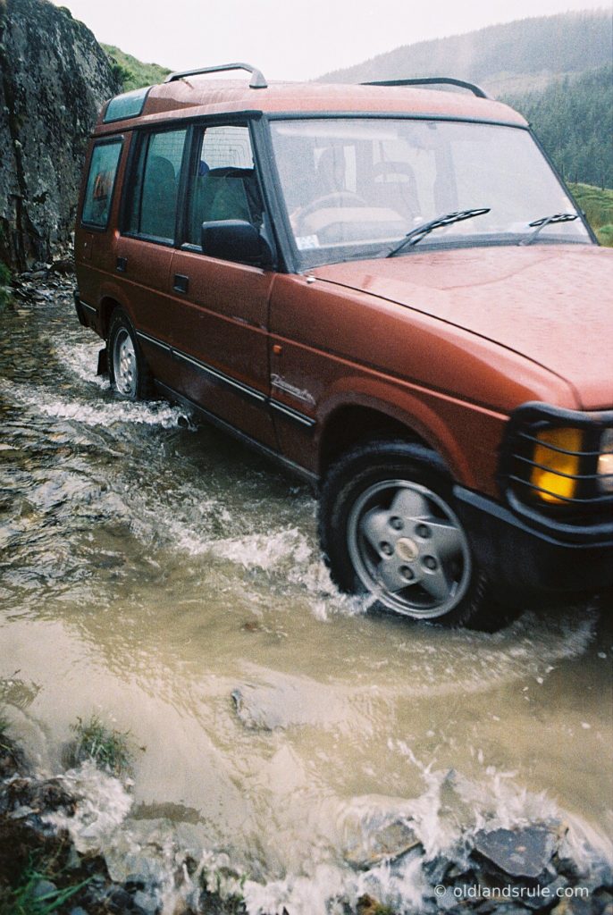 A red Land Rover V8N Discovery driving through a shallow stream, making small waves. The front of the car is just out of shot but you can see the edge of the bull bars and that the headlamps are on. There is big rock, trees and a hill in the background