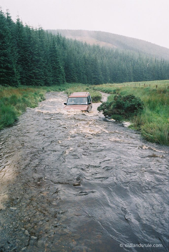 Red Land Rover V8 wading through a stream with the water washing over the bonnet and fir trees and a big hill in the background