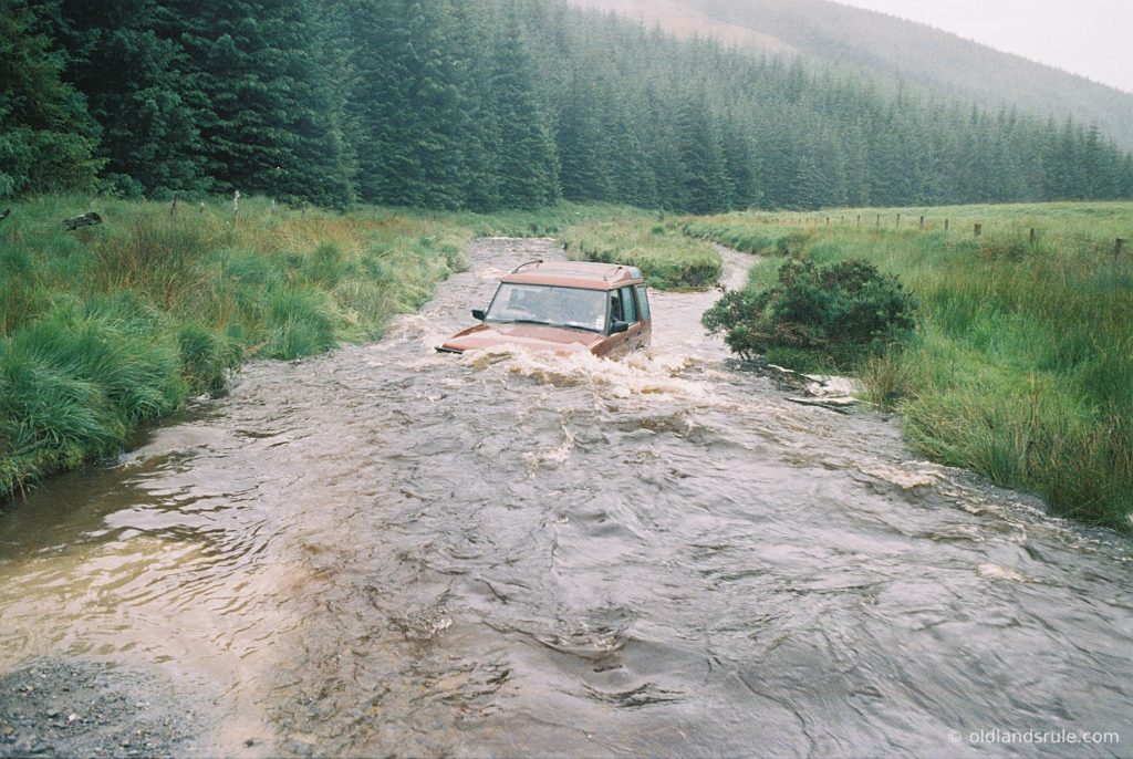 A red Land Rover Discovery wading a river at Strata Florida, Wales. There are pine trees in the background and the water is washing up and over the bonnet of the car