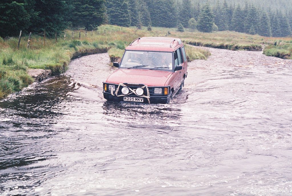 A red Land Rover Discovery 1 bumper deep in the middle of a small river with fir trees in the background. There's a recovery rope wound around the bullbars.