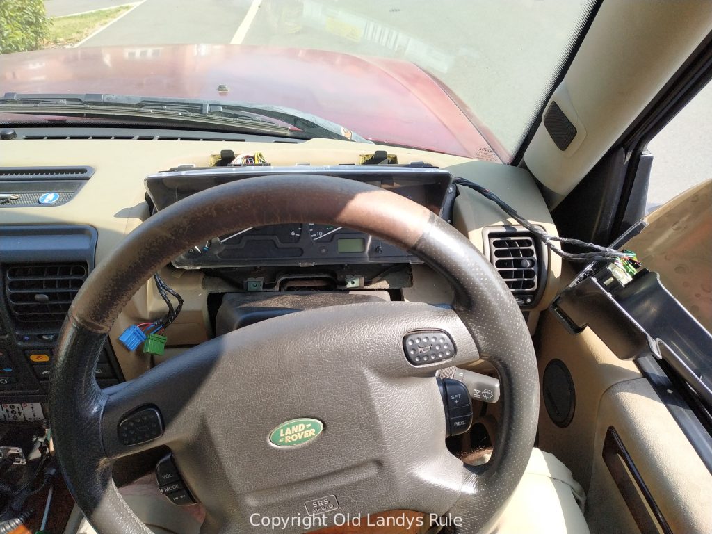 Looking down on a Discovery 2 steering wheel, with the dashboard surround removed and hanging out of the side window still attached by it's cables.