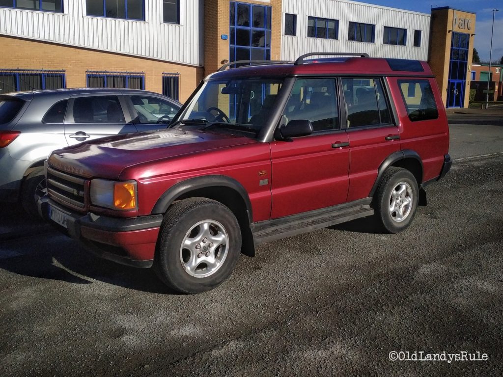 Metallic red Discovery 2, side view, in a car park with a commercial building behind it.