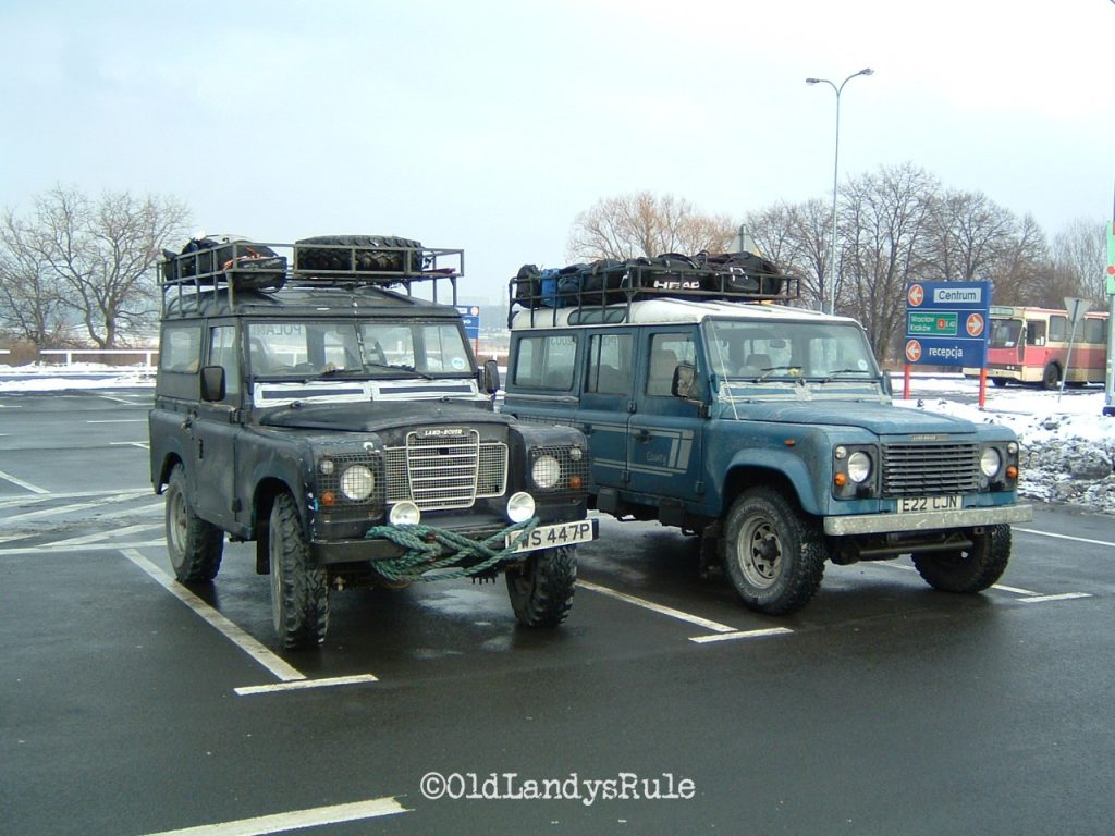 Two Land Rovers, side by side, in a car park in Poland. A blue Land Rover Series 3, with a roofrack, spotlights, rope looped around the bumper and silver gaffer tape over the front vents. And a blue One-ten County, with a roof rack and silver gaffer tape around the front vents