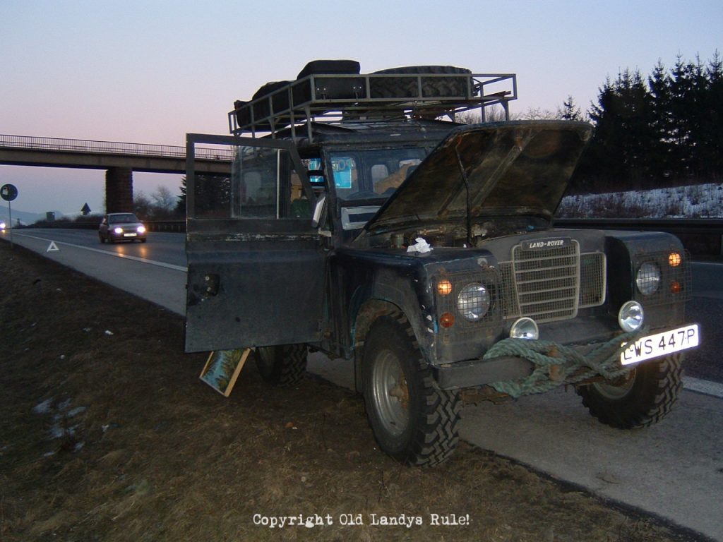 A blue Land Rover Series 3, parked on the hard shoulder of a German autobahn at dusk, with its drivers door open and the bonnet up.