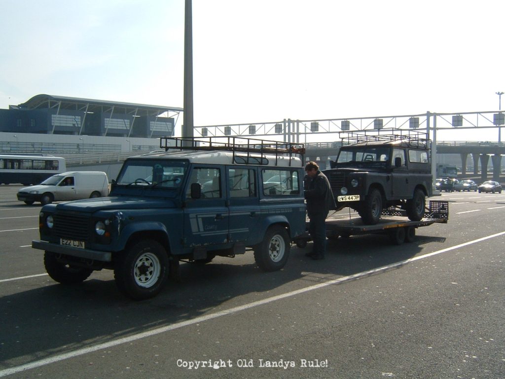 A blue Land Rover One-ten, stationary at Calais docks waiting to load on the ferry. A trailer is attached and there's a blue Land Rover Series 3 on it.