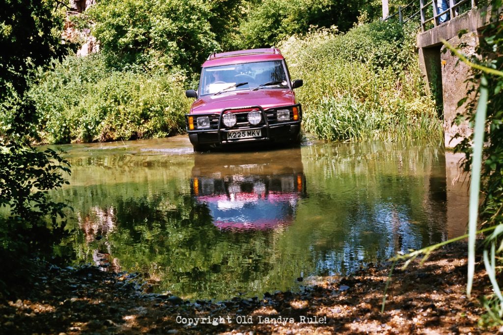Red Land Rover Discovery 1, approaching the camera, through a river. There is a bridge to the right and greenery all around