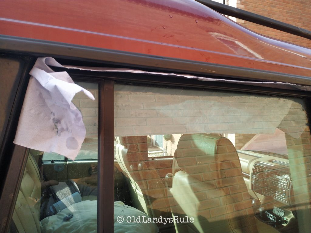 Looking through the rear right side window of a red Land Rover Discovery 2, there is blue paper towel tucked in the top of the door and can be seen hanging down inside the car
