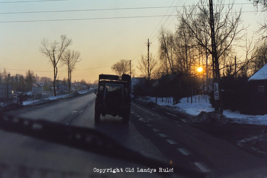 Taken through the windscreen of a Land Rover, capturing another Land Rover in front, on a Polish road, at dusk. The sun is shining through the trees at the side of the road and there are small buildings covered in snow.