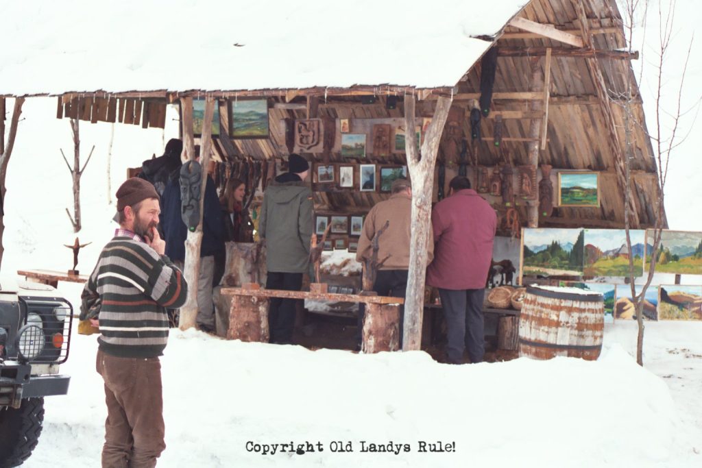 An open sided wooden hut, selling paintings, set in snow. There's a man in the foreground, and the corner of a Land Rover One-ten is just visible