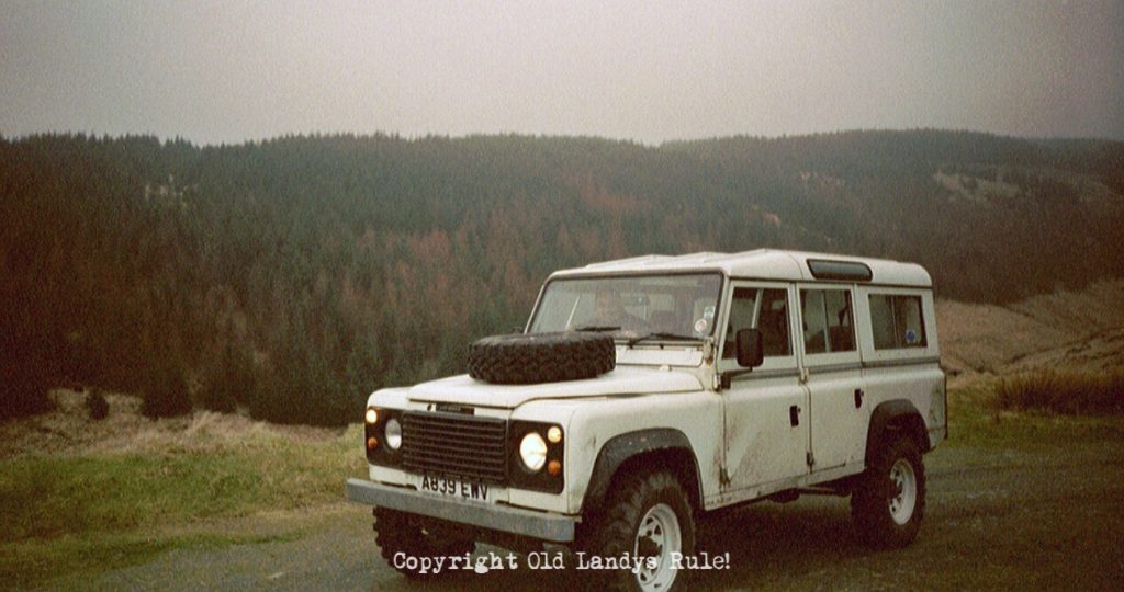 A white 1984 Land Rover 110 V8 with a Welsh pine forest as the backdrop