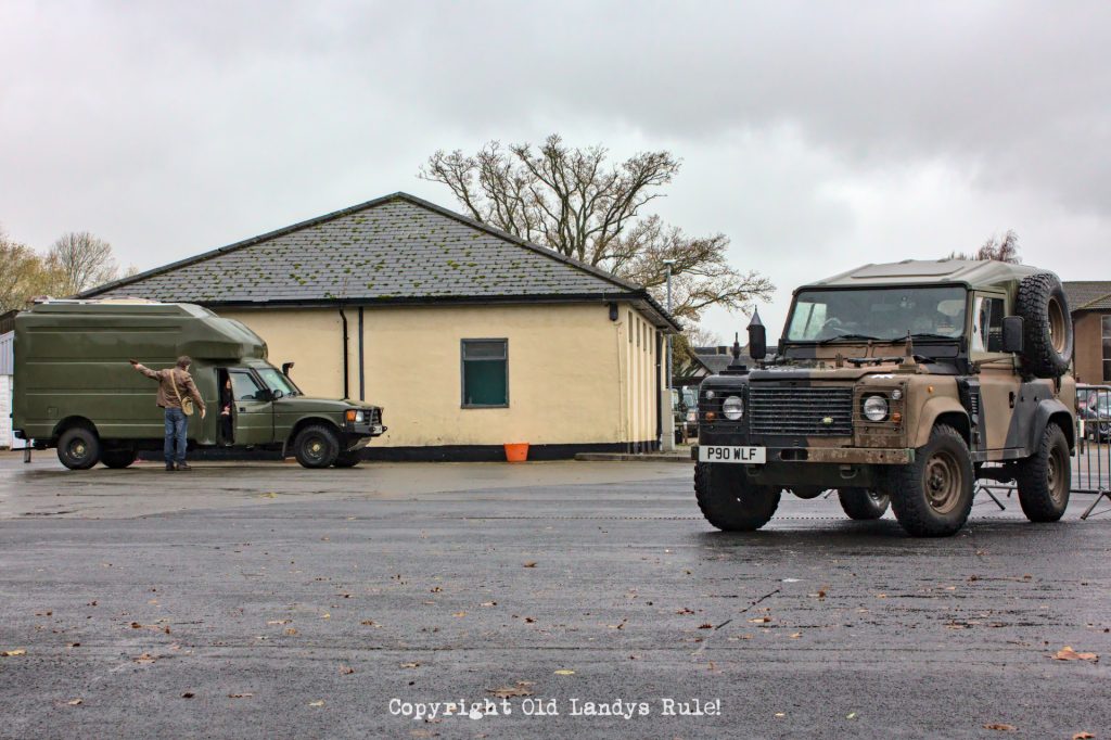 Looking across a carpark at a Land Rover Ninety 'Wolf' on the right side and a Land Rover Discovery 1 with what appears to be a custom camper body. The Ninety is in camo colour and the Discovery 1 is dark green.