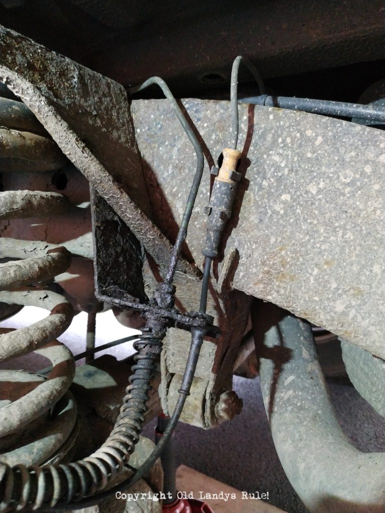 Close-up of an old, rusty, and leaking brake pipe attached to a flexi-hose. There is a suspension coil spring just visible to the left