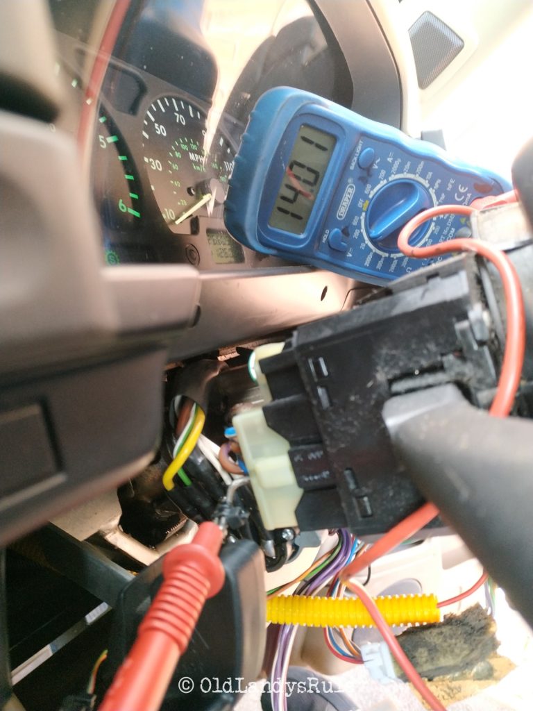 Closeup of a multimeter with 14.01 volts reading on the display. There's some dashboard clocks visible in the background