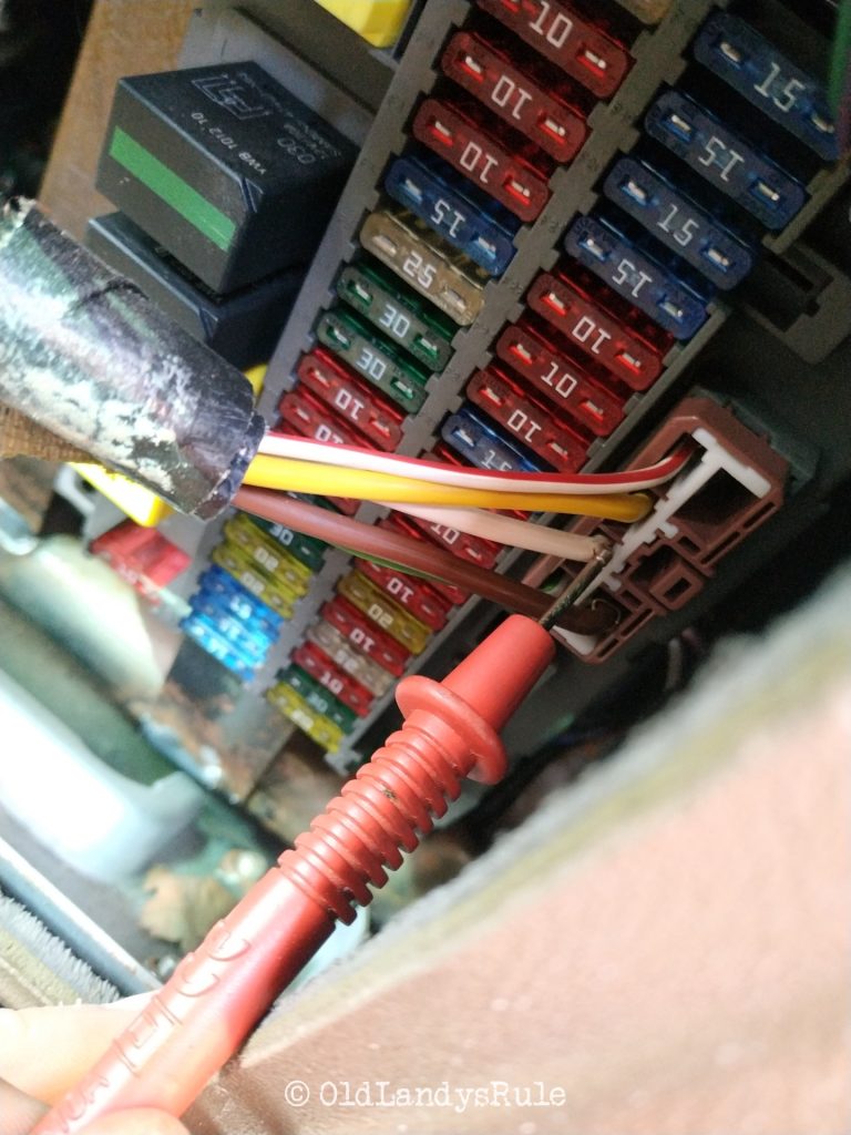 A multimeter probe, touching a specific wire in a connector block, that's inserted into a car fusebox